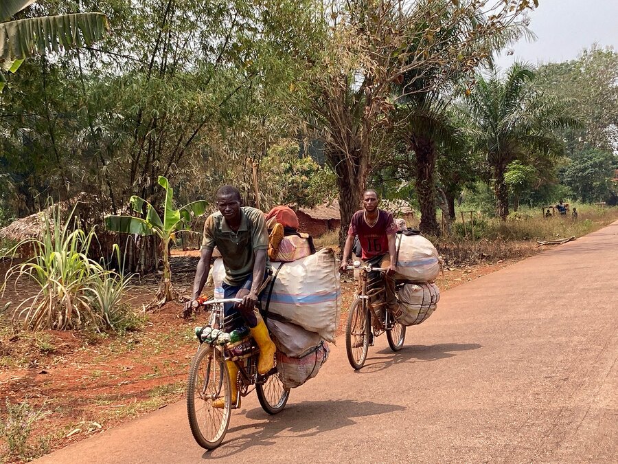 Two men cycle along a road and their bicycles carry large bags of food to sell at the market.